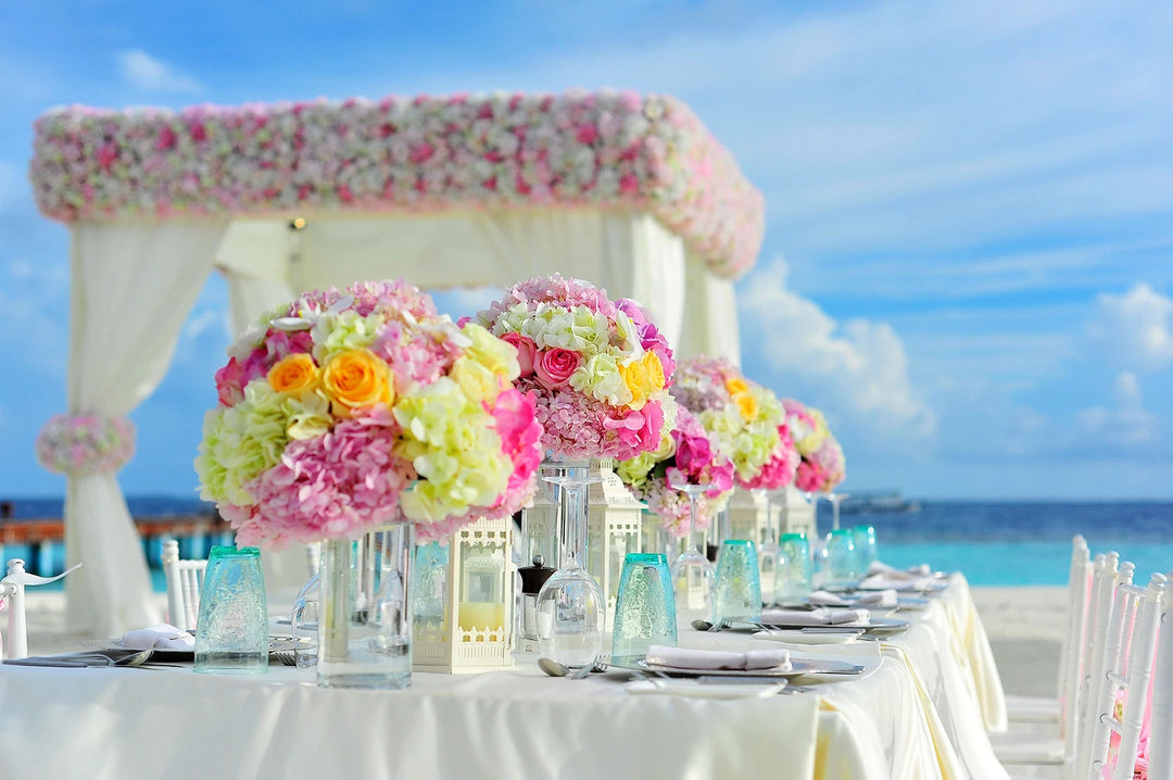   A table top with glasses, plate setting and colorful flower vase in the centre set outdoors near the ocean for a wedding party